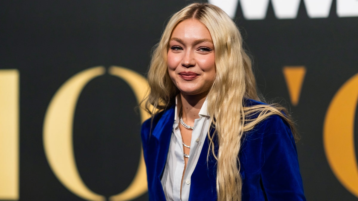 Gigi Hadid in a white button down top and dark blue, shiny velvet blazer jacket soft smiles at the camera