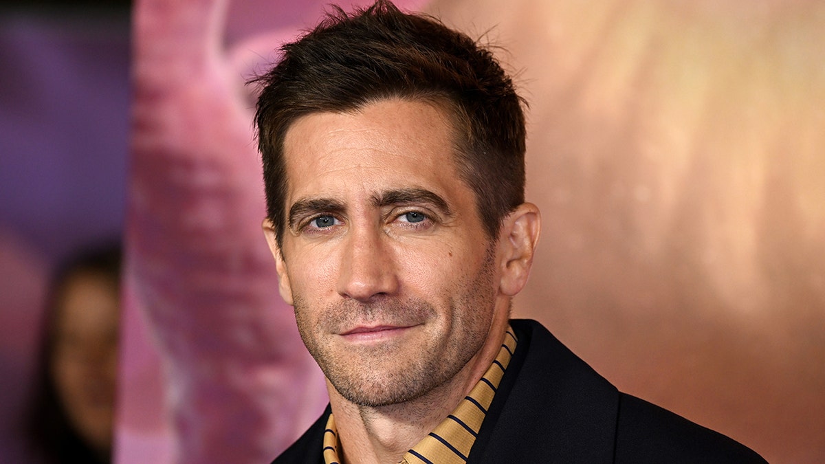 Jake Gyllenhaal in an orange striped shirt and a black jacket on the red carpet smiles softly