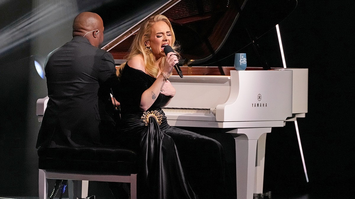 Adele sits on a piano seat and sings into the microphone wearing a black velvet dress while a man in a black suit plays a white piano on stage in Vegas