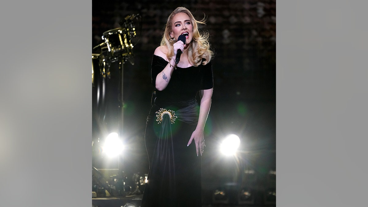 Adele stands on stage singing into her microphone in a black gown with a gold clasp holding a sash of her dress in Vegas