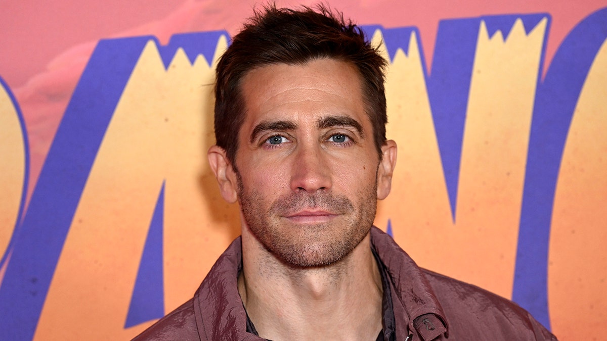 Jake Gyllenhaal looks directly at the camera in a purple/red jacket on the red carpet for the "Strange World" multimedia event