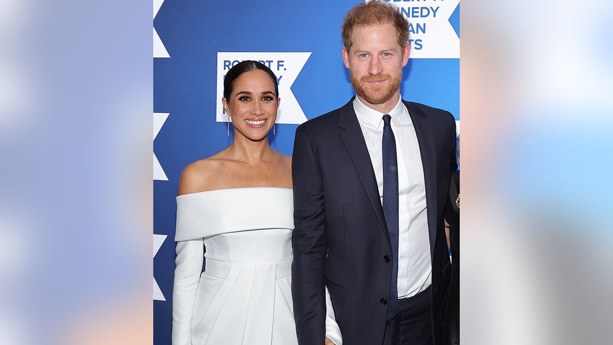 Meghan Markle in a white off the shoulder long sleeve gown holds hands with Prince Harry in a navy suit and tie at the Robert F. Kennedy Human Rights Ripple of Hope Gala red carpet