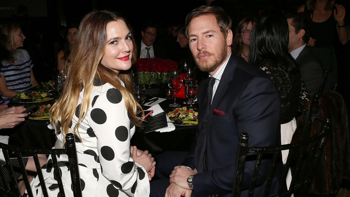Drew Barrymore in a white dress with black polka dots is seated next to husband Will Kopelman at a table