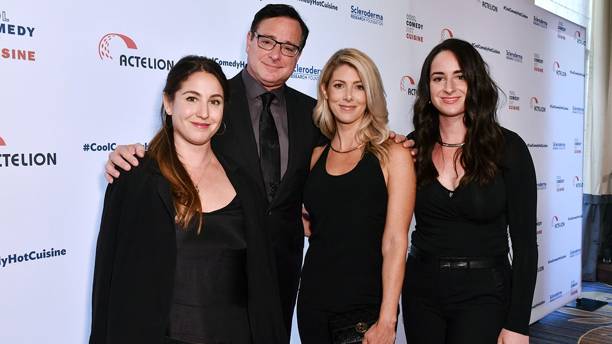 Aubrey Saget in all black stands next to father Bob Saget in a black suit and tie, next to wife Kelly Rizzo in a black dress, next to Lara Saget, also in all black on the red carpet
