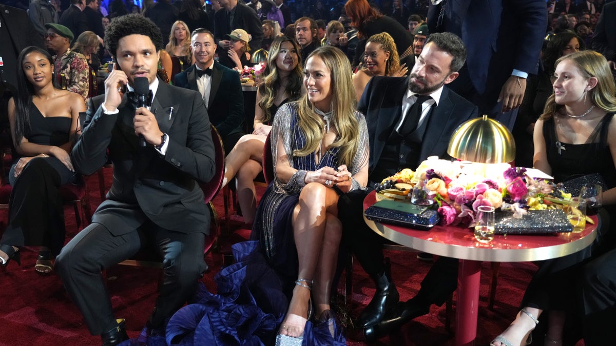 Trevor Noah on a phone at the Grammy table of Jennifer Lopez, wearing blue, and Ben Affleck, wearing a suit.