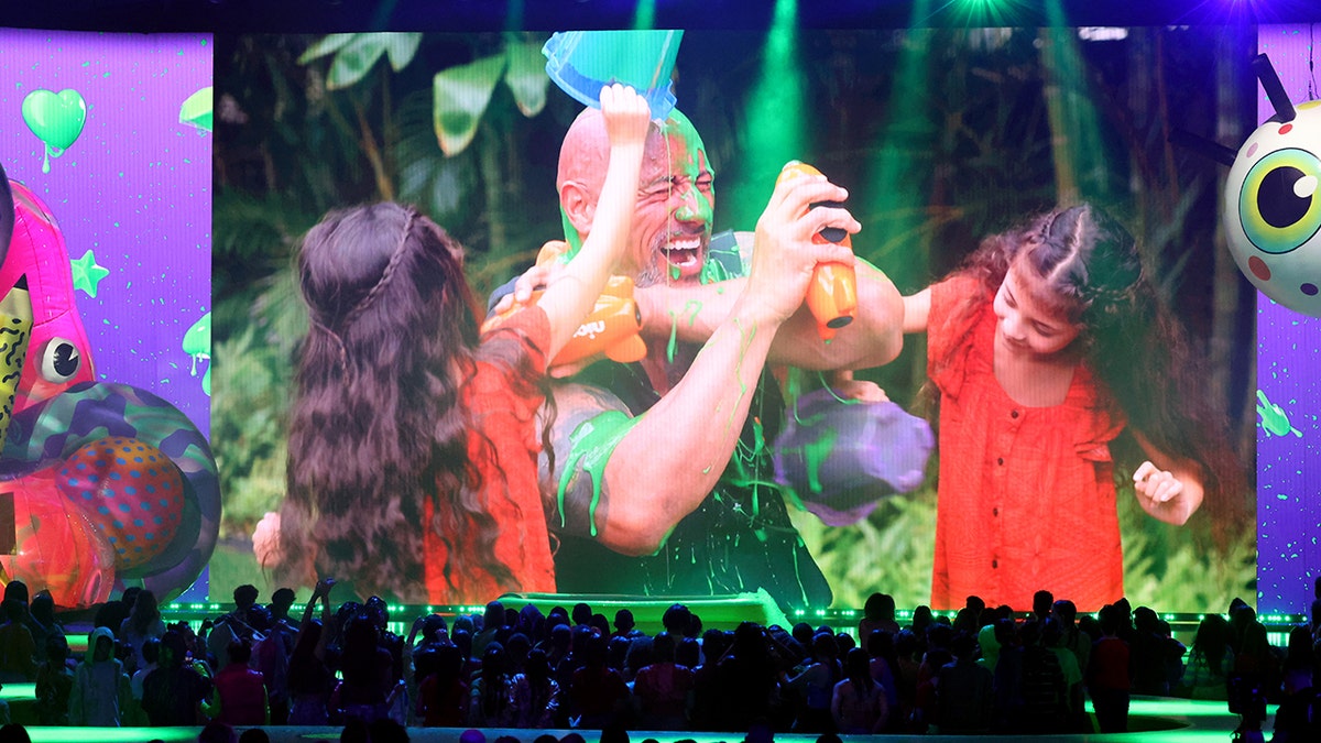 Dwayne Johnson is covered in slime as his daughters Jasmine and Tia empty buckets of green slime on him while he appears on a big screen at the Kids' Choice Awards