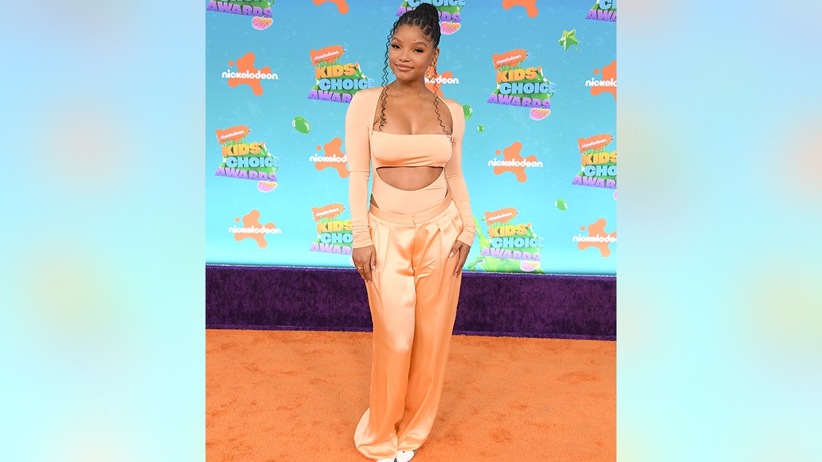Halle Bailey wears an orange bodysuit and matching satin pants on the orange carpet at the Kids' Choice Awards
