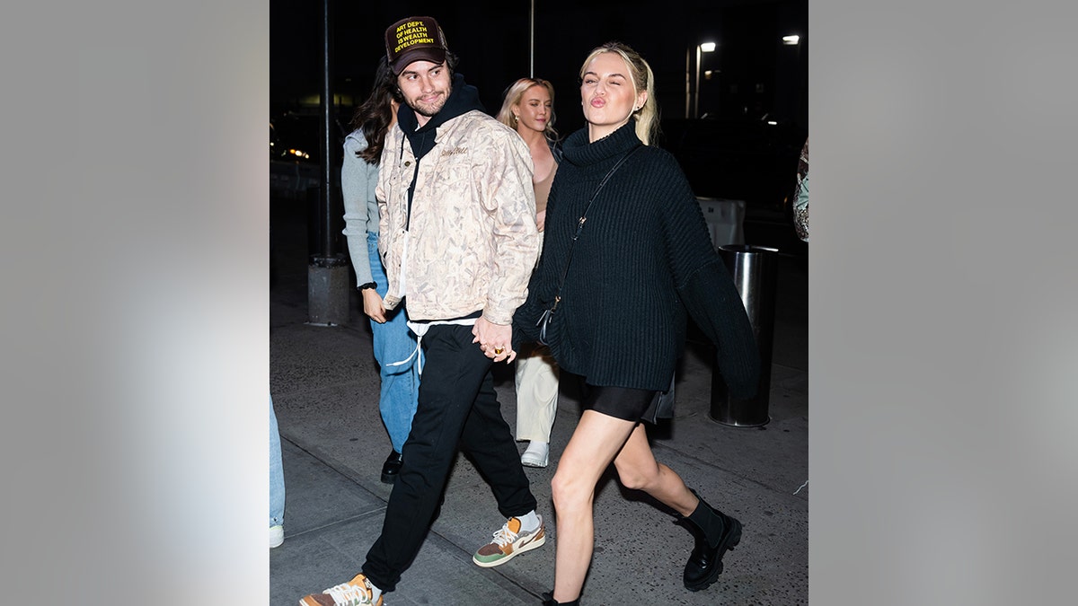 Chase Stokes in a black hoody and pants with a patterned jacket on and brown hat holds hands and smiles at Kelsea Ballerini in black biker shorts and a black sweater giving a kissy face to the camera
