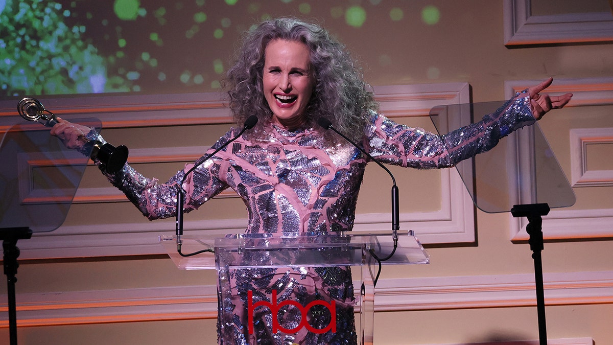 Andie MacDowell on stage at the Hollywood Beauty Awards accepting her award for Timeless Beauty, with her arms spread out in a pink and silver dress