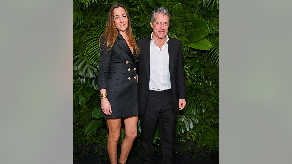 Anna Elisabet Eberstein in a black suit dress with large gold buttons poses with husband Hugh Grant in a white button down and black suit at the Chanel and Charles Finch dinner