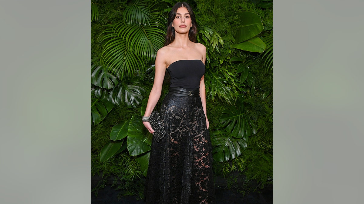 Camila Morrone in a long black dress with a lace bottom holds a sparkly Chanel bag and unique Chanel cuff at the Chanel and Charles Finch dinner