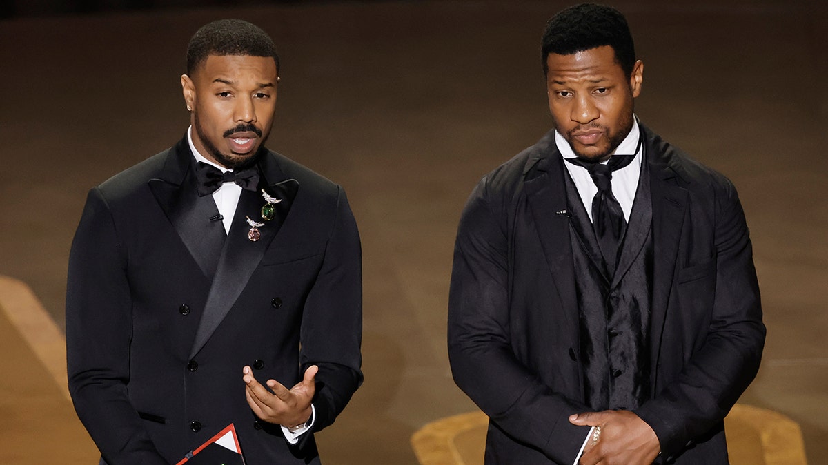 Michael B. Jordan in a black tuxedo speaks on stage while he presents the award for best cinematography with Jonathan Majors, in a multi-piece suit