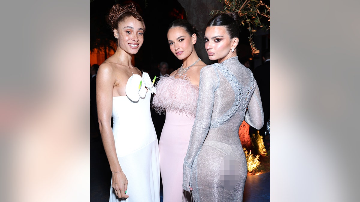 Adwoa Aboah in a white gown with two orchids at her bust stands next to Lily James in a pink dress with a feathery top next to Emily Ratajkowski who shows her butt in a completely sheer dress