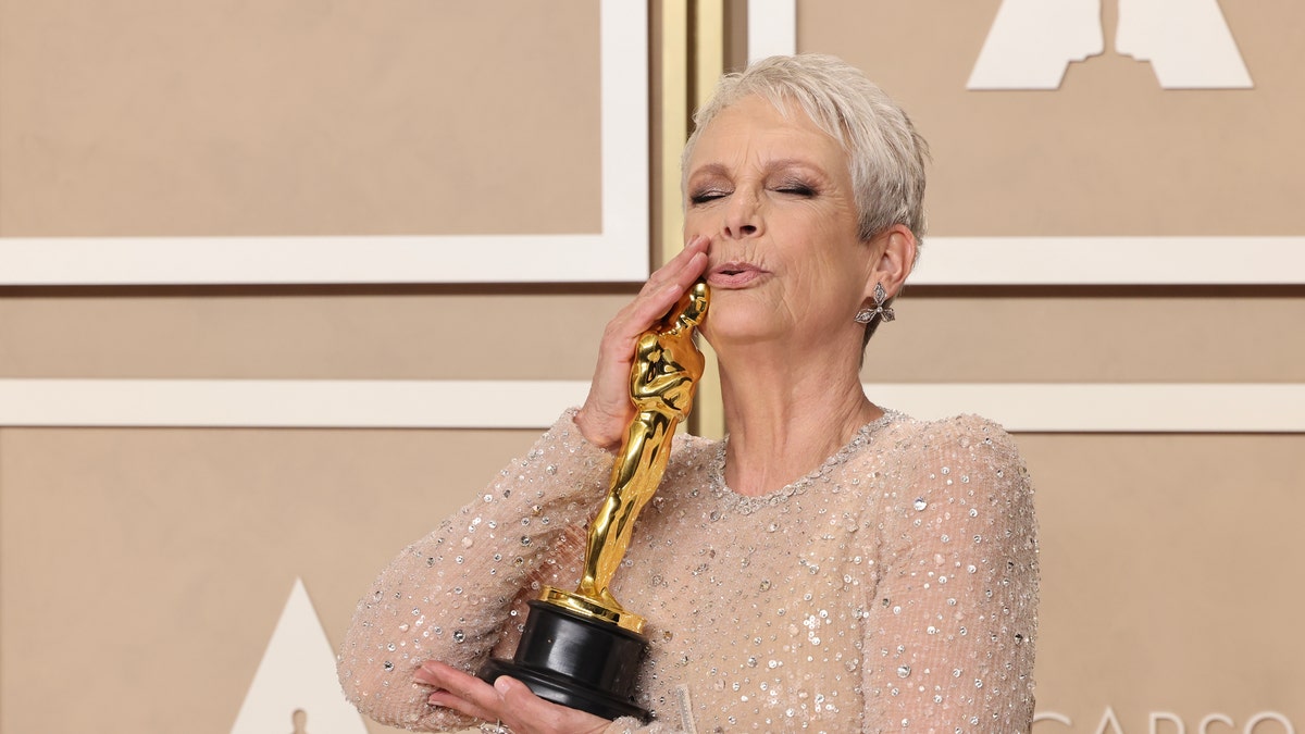 Jamie Lee Curtis in her sheer white dress with sparkles holds her Oscar award up to her face and kisses the head of the Oscar