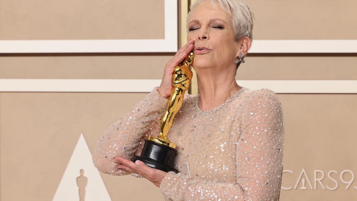 Jamie Lee Curtis in her sheer white dress with sparkles holds her Oscar award up to her face and kisses the head of the Oscar