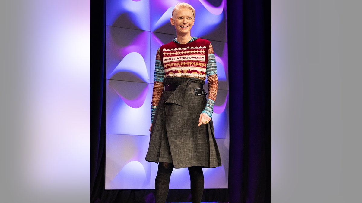 Tilda Swinton walks on stage in a long black skirt, black tights, a colorful long-sleeve shirt and red vest