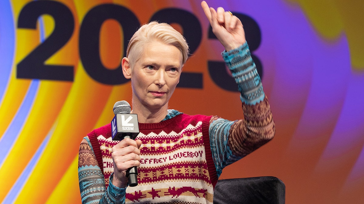 Tilda Swinton shakes her finger on the SXSW stage wearing a colorful top and red knit vest
