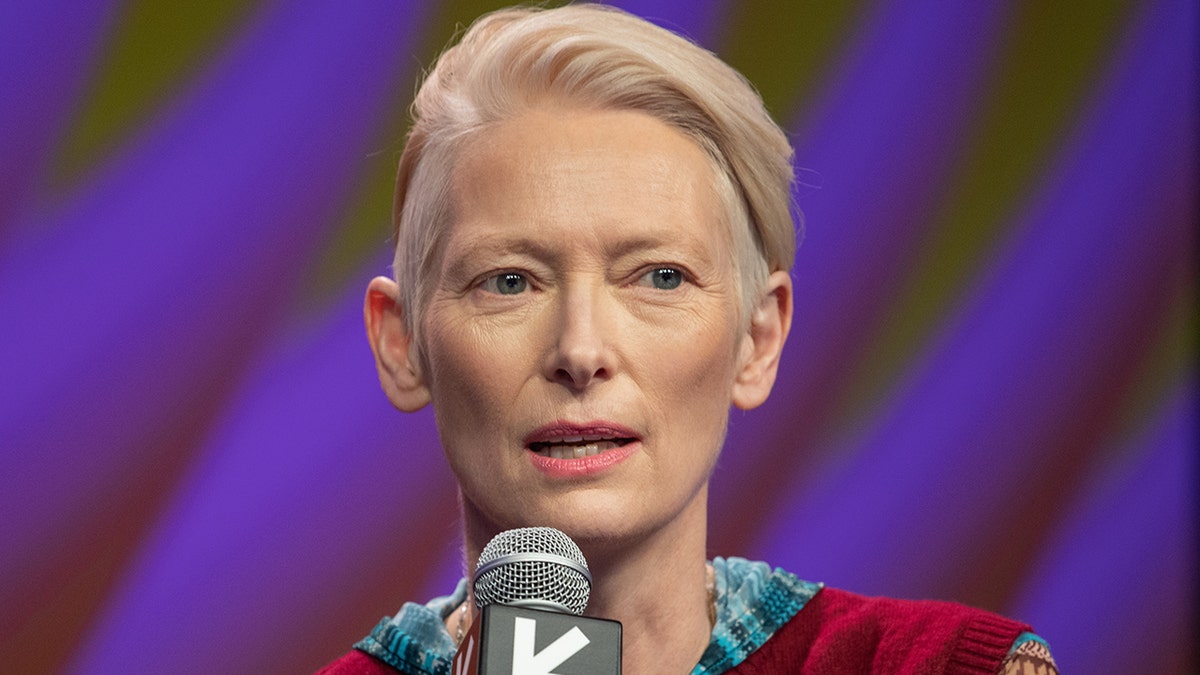 Tilda Swinton speaks into a microphone at SXSW as a keynote speaker in a red vest and multi-colored long-sleeve shirt