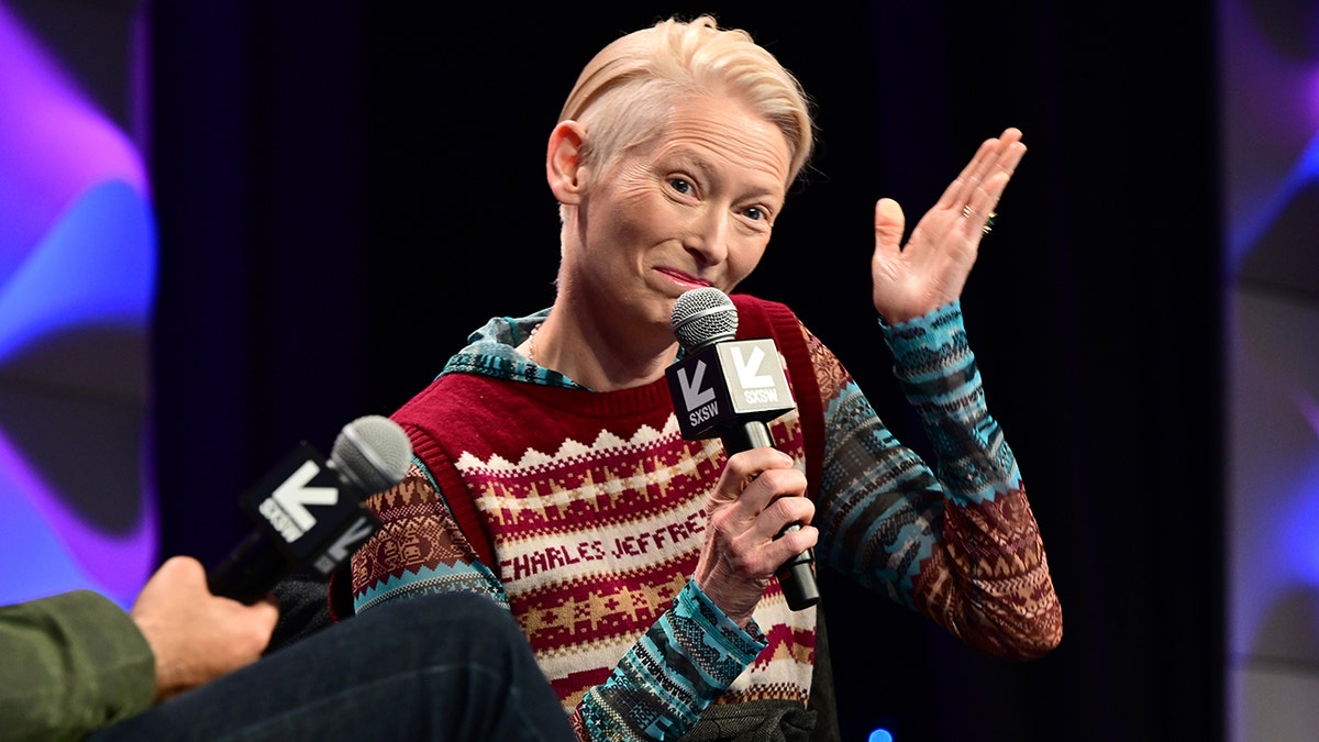 Tilda Swinton shrugs on stage while holding a microphone at SXSW in a colorful long-sleeve shirt and red vest