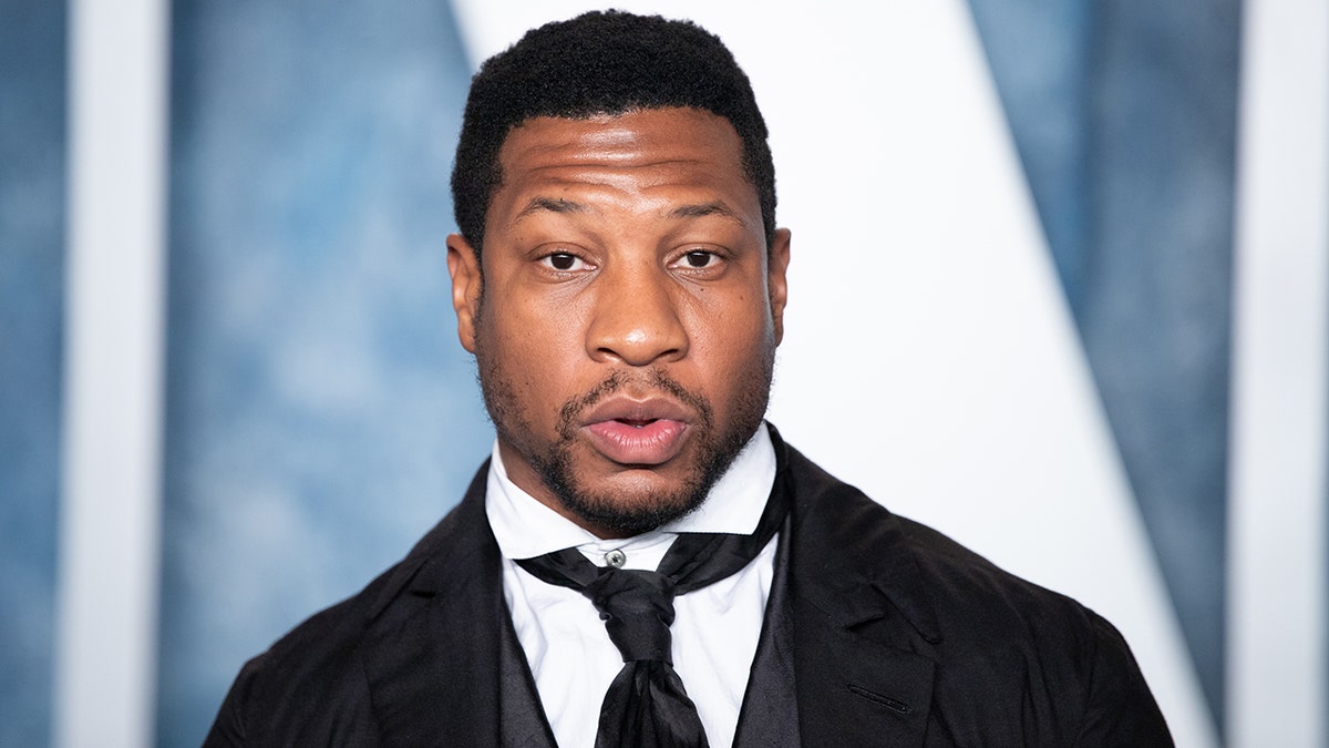 Jonathan Majors looks directly at the camera wearing a black suit, white shirt and slightly loose black tie at the Vanity Fair Oscar party