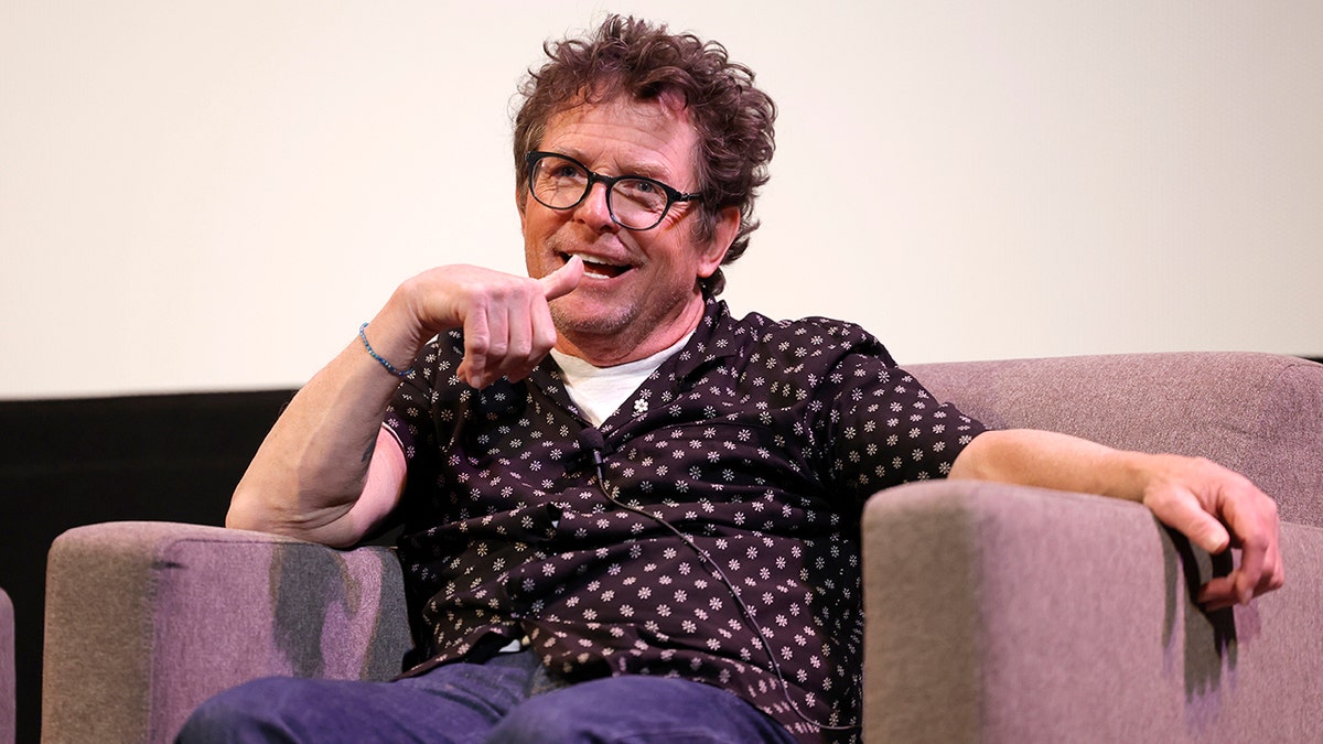 Michael J. Fox sits in a tan chair on the SXSW stage, in a black top with white flowers and jeans, wearing black rimmed glasses