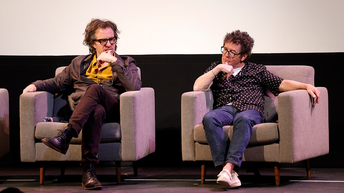 David Guggenheim in a mustard shirt and brown jacket, pants, boots and frames sits on a tan chair beside Michael J. Fox in a black shirt with white flowers on it, while on stage for a Q+A at SXSW