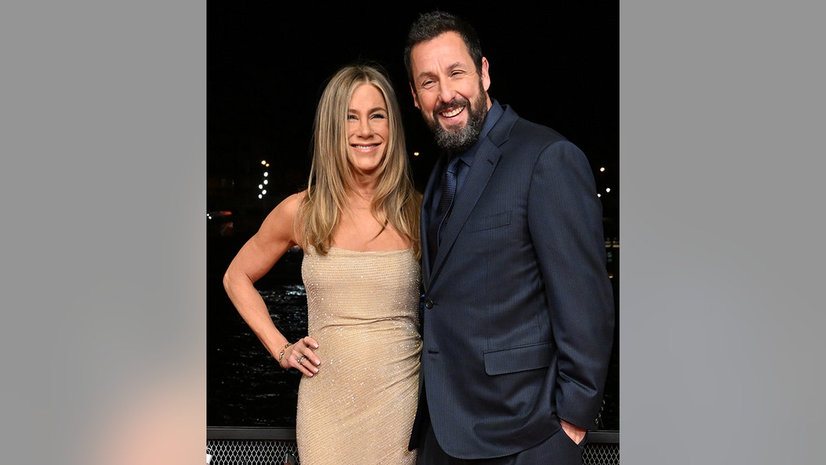 Jennifer Aniston in a sequined gold dress puts her right hand on her hip and smiles for a photo with Adam Sandler wearing a navy suit and tie