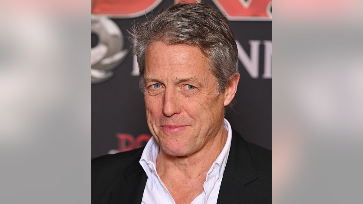 Hugh Grant wears a light blue shirt and black jacket on the red carpet in Paris France and soft smile/smirks for camera