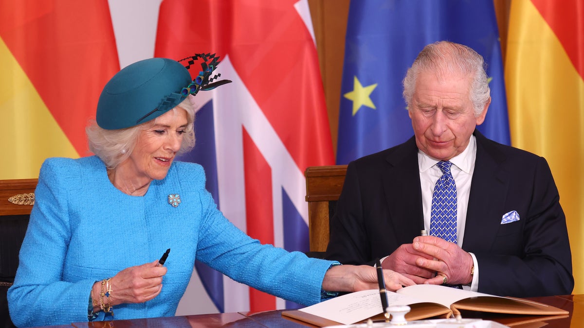 Camilla in a teal coat and turquoise hat reaches over to sign a book during a reception in Germany as King Charles with a bright blue tie and black jacket watches