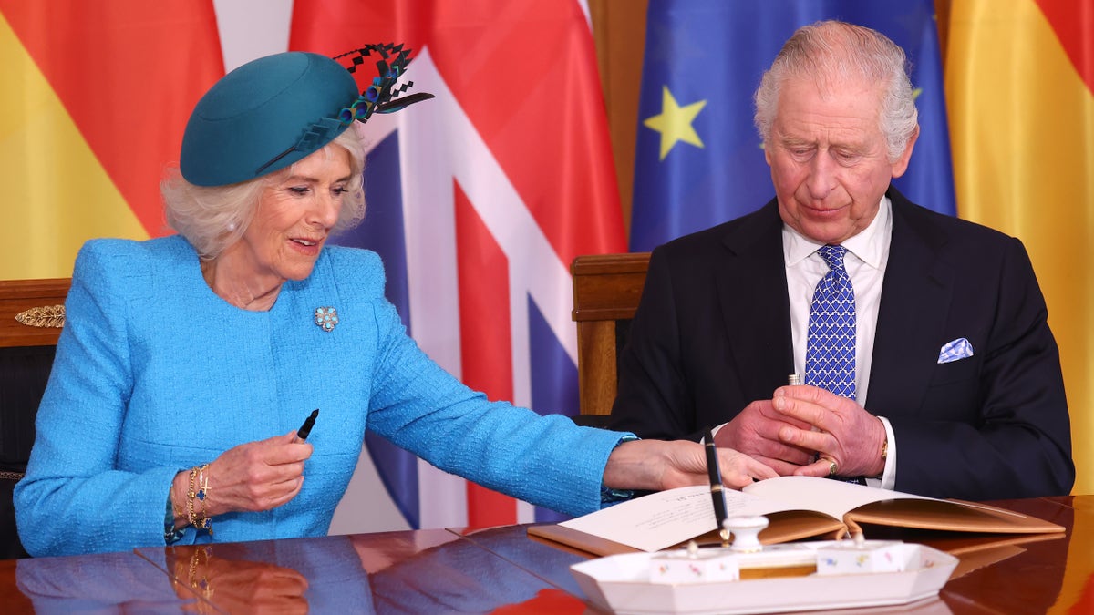 Camilla in a teal coat and turquoise hat reaches over to sign a book during a reception in Germany as King Charles with a bright blue tie and black jacket watches