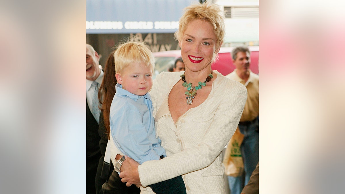 Sharon Stone in a white sweater and green necklace holds her son Roan wearing a blue button-down shirt on the red carpet