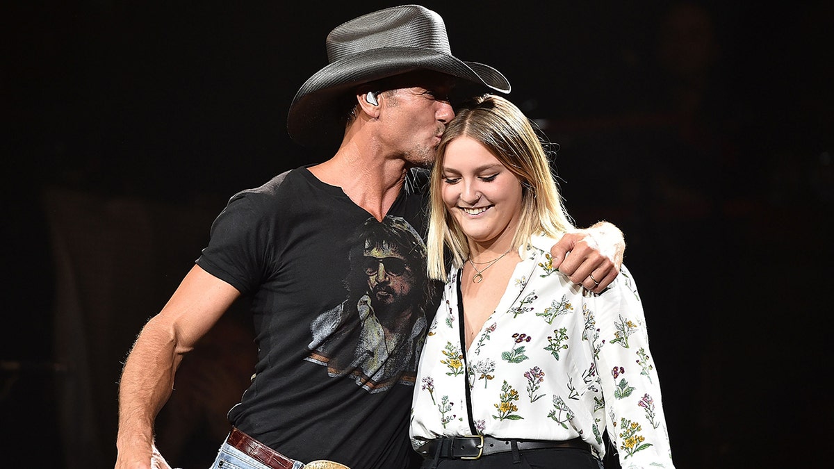 Tim McGraw with a black shirt and cowboy hat kisses daughter Gracie's head in a white printed blouse on stage in Nashville