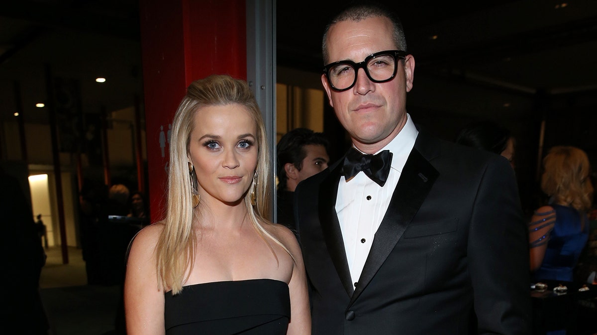 Reese Witherspoon in a strapless black dress stands next to husband Jim Toth in a black tuxedo and dark thick rimmed black glasses