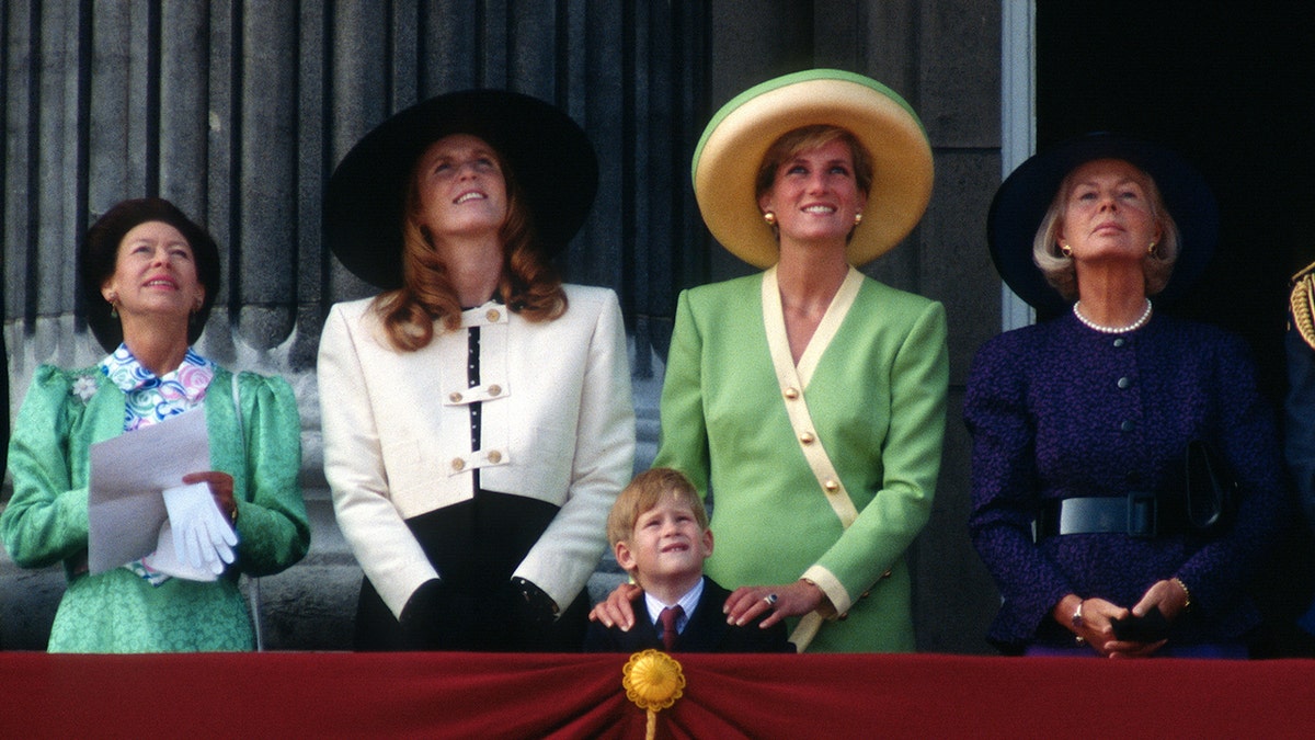 Princess Margaret wears an emerald jacket with a multi-colored shirt and looks up next to Sarah Ferguson, Duchess of York wearing a white cropped jacket and a black hat, next to Diana, Princess of Wales in a green dress and hat with yellow trim, holds on to the shoulders of Prince Harry in a suit and red tie next to Katharine, Duchess of Kent in a navy outfit and hat