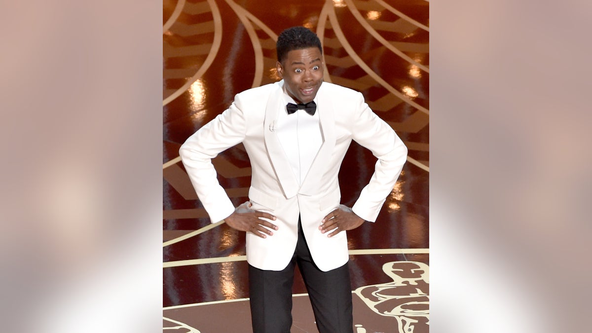 Chris Rock puts his hands on his hips wearing a white tuxedo and black pants with a black bowtie while hosting the Academy Awards in 2016