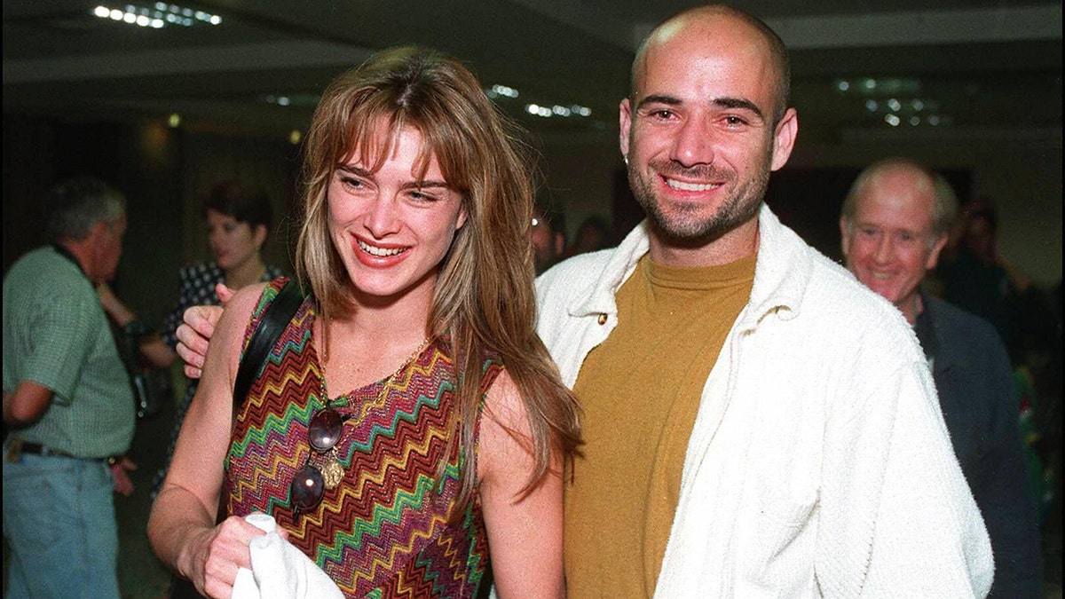 Brooke Shields in a zig-zag tank top smiles in front of Andre Agassi wearing a mustard shirt and white jacket in Cape Town