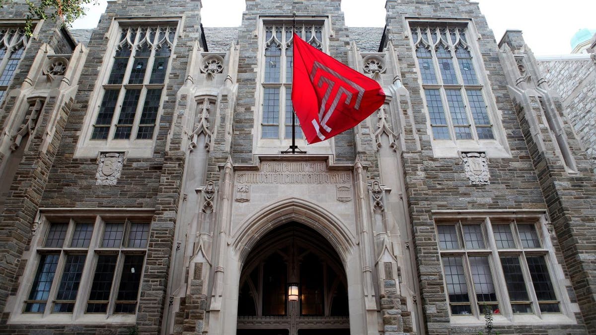 Gray Temple University building exterior with rows of large windows and the university flag hanging above an arched doorway