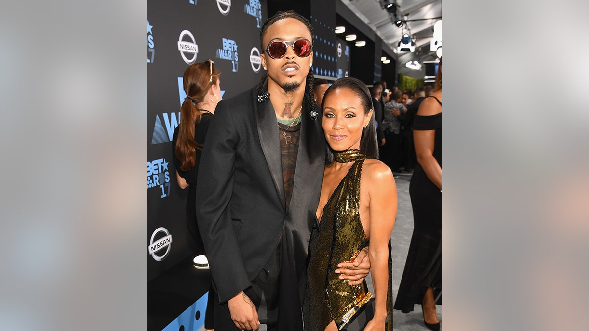 August Alsina in a black suit and sunglasses poses with Jada Pinkett Smith in a black cut-out outfit on the red carpet for the BET Awards