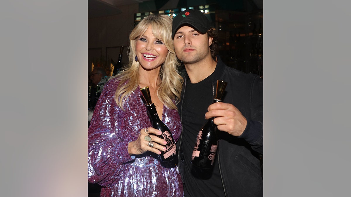 Christie Brinkley in a sparkly purple dress holds a bottle next to son Jack in a black outfit and black hat, also holding a bottle