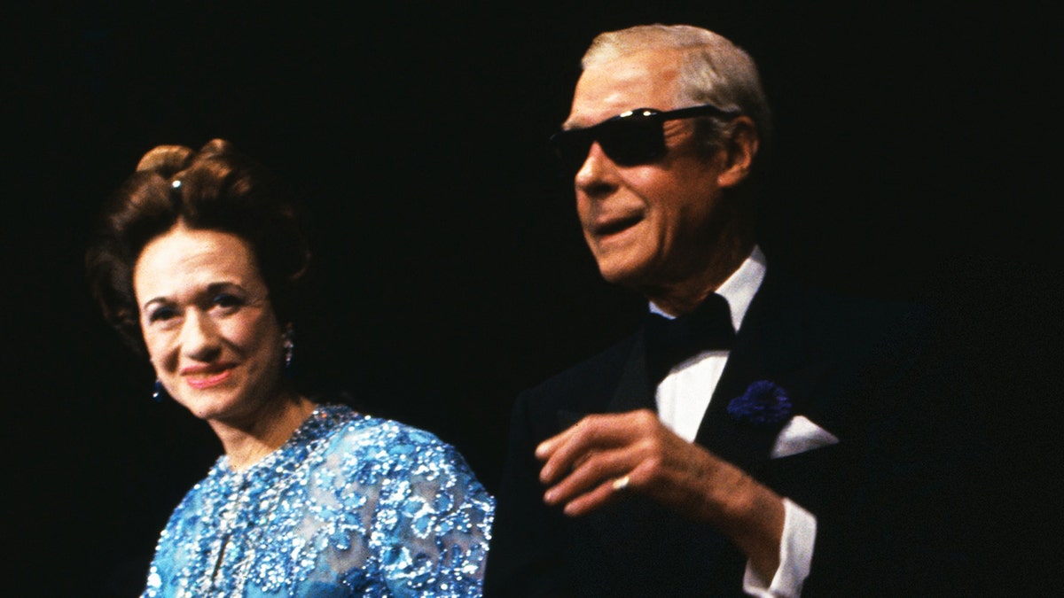 Wallis Simpson wearing a bright blue dress while standing next to her husband Edward VIII wearing a tux