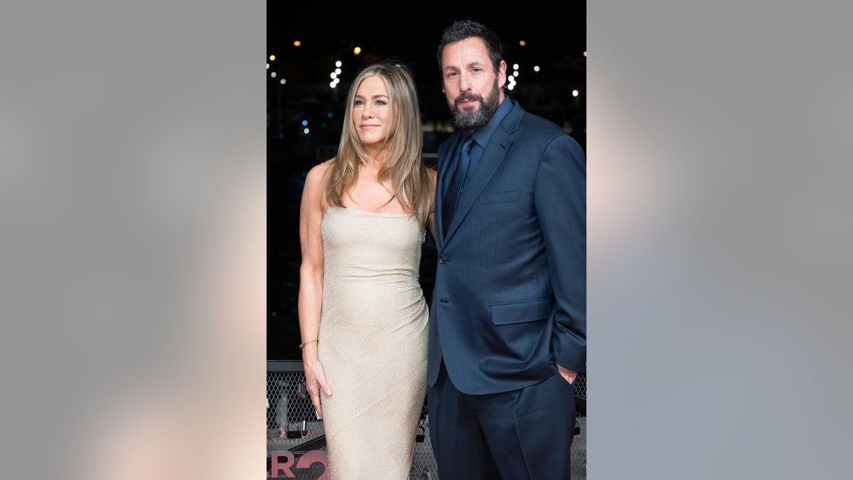 Adam Sandler wearing a dark blue suit next to Jennifer Aniston wearing a nude hued dress