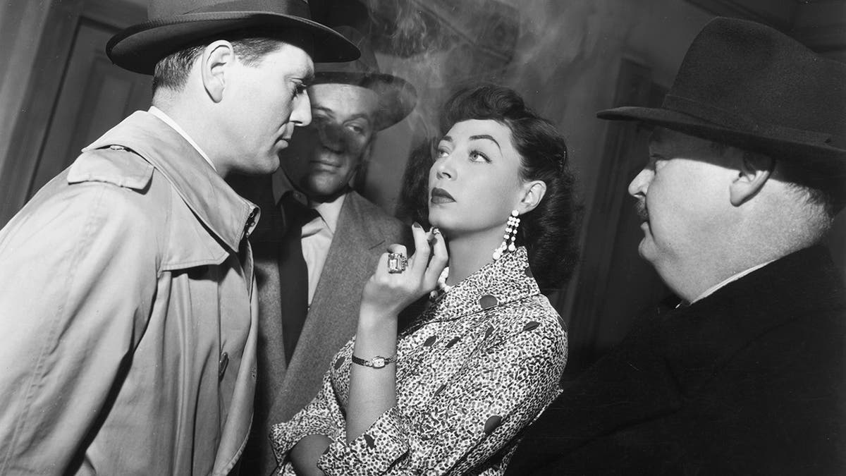 Marie Windsor in a film scene surrounded by actors playing detectives while she smokes