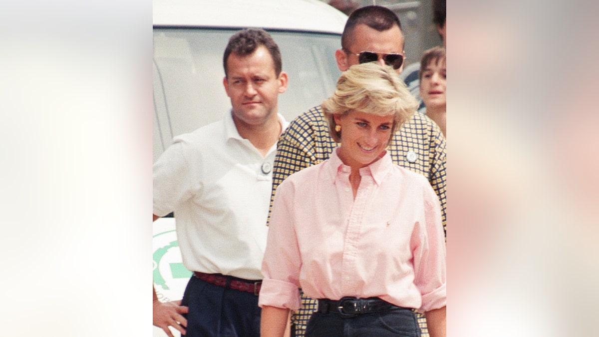 Paul Burrell in the background wearing a white shirt with Princess Diana up close and smiling in a pink blouse and dark pants