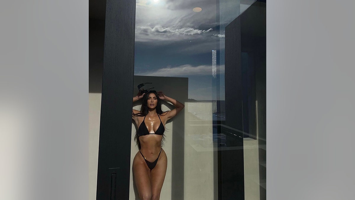 Kim Kardashian shows off her hour-glass figure in a black bikini with her hands behind her head as she stands behind a sliding glass door