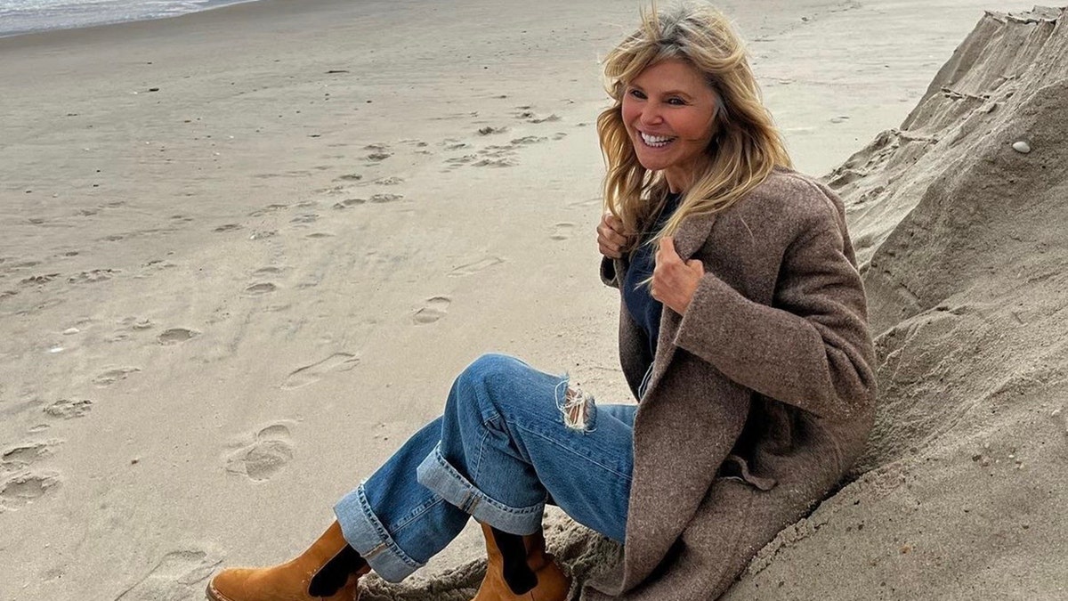 Christie Brinkley in a cozy sweater and ripped jeans smiles while sitting in the sand at the beach showing off her gray roots