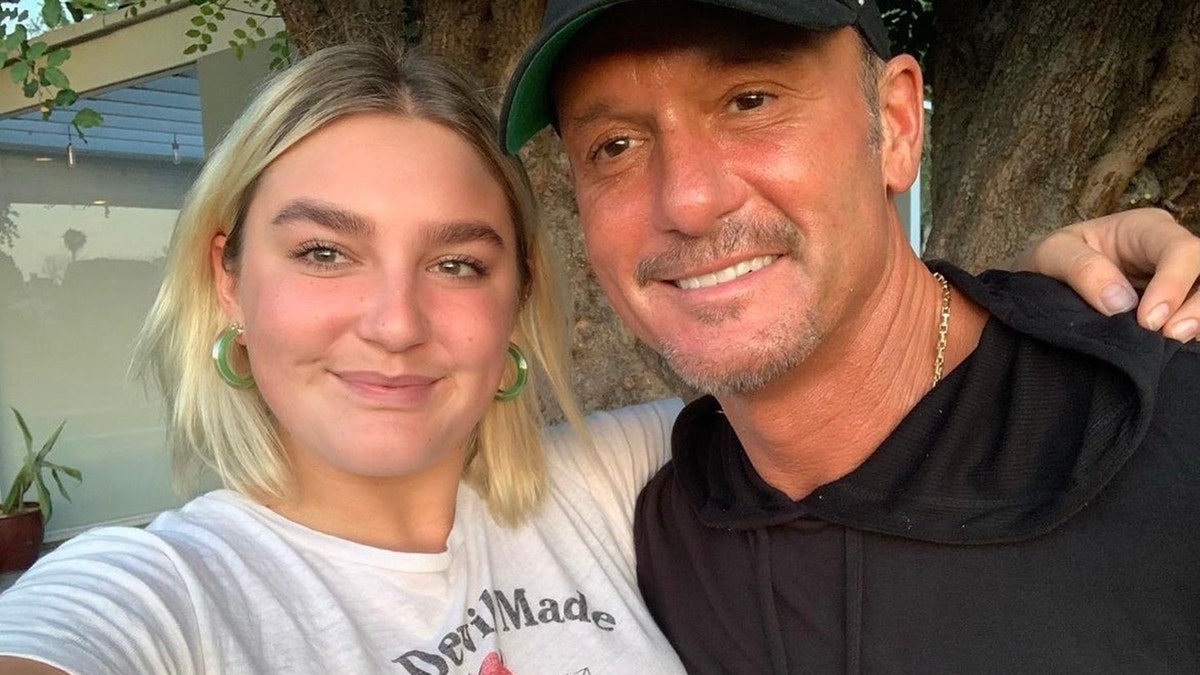Tim McGraw in a black shirt and hat smiles with daughter Gracie wearing a white t-shirt