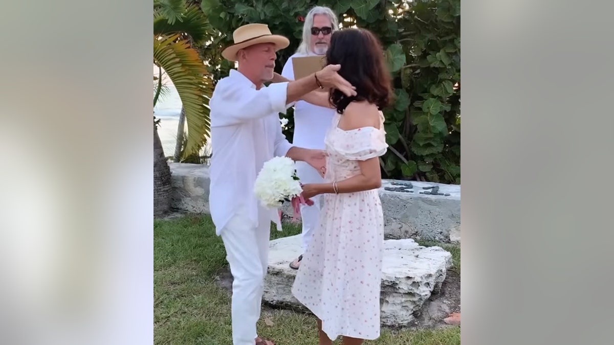 Bruce Willis in a white shirt and pants moves to give his bride Emma in a white dress with a tiny pattern and long ruffle top a kiss during their vow renewal in Turks and Caicos in 2019