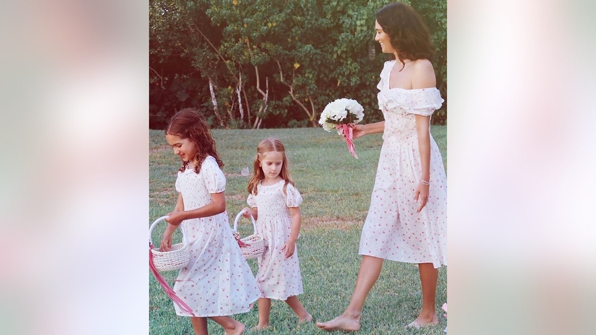 Emma Heming holds a bouquet of off-white flowers wearing a white dress with a tiny pattern, walking in front of her with matching dresses are daughters and flower girls Mabel Ray and Evelyn Penn