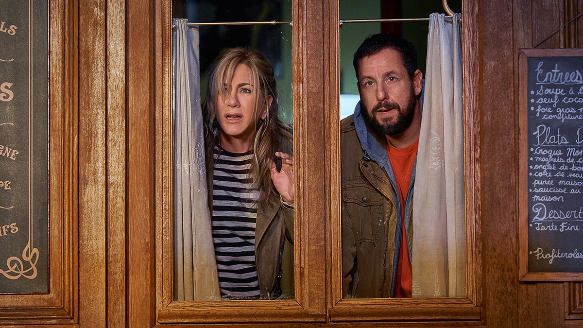 Still photo of Jennifer Aniston as Audrey Spitz and Adam Sandler as Nick Spitz in "Murder Mystery 2" looking out a restaurant window, looking confused