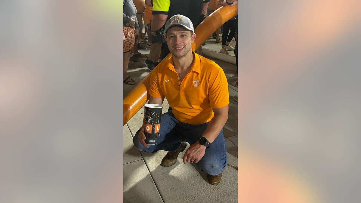 Collazo crouches down holding a cup of soda, wearing jeans and an orange Tennessee Volunteers polo shirt and cap